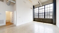 This single-floor loft showcases a compelling blend of industrial character and modern finishes throughout. The main living area is defined by dramatically high ceilings with exposed ductwork, polished concrete floors, and a striking wall of large, steel-framed factory-style windows set above a dark brick base — a nod to the building's industrial heritage. Track lighting along the ceiling complements the abundant natural light that pours in through the oversized windows.

The kitchen is well-appointed with two-tone cabinetry featuring gray lower cabinets and white upper cabinets, a light tile backsplash, white countertops, and a full suite of stainless steel appliances including a refrigerator, range, and over-the-range microwave. Open shelving adds functional storage and visual interest.

The bedroom continues the high-ceiling aesthetic with exposed ductwork overhead, polished concrete flooring, and a dedicated closet with built-in shelving. The bathroom is clean and contemporary, featuring a combined tub and shower with a frameless glass sliding door, a streamlined white vanity, and stone-textured tile flooring. The overall ambiance is open, airy, and distinctly urban.