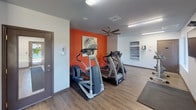 This single-story fitness center is designed to provide a comprehensive workout experience in a clean, modern environment. The space is defined by a bold color palette, with striking orange accent walls contrasting against neutral white and gray tones, giving the facility an energetic and motivating atmosphere. Durable wood-look flooring runs through the main areas, while carpeted flooring in the strength training zone adds a practical touch underfoot.

The cardio area is well-appointed with a selection of treadmills, elliptical trainers, and an upright stationary bike, all positioned to make use of the natural light filtering in through multiple windows. A ceiling fan overhead contributes to comfort during workouts. Wall-mounted artwork adds a finishing aesthetic touch to the space.

A dedicated strength training zone features a multi-station cable machine, a weight bench, and a full rack of dumbbells, catering to a wide range of resistance training needs. A wall-mounted fan provides additional ventilation in this section.

The entrance area is bright and welcoming, with large glass doors that offer views of the surrounding outdoor greenery, a window with frosted signage, and a clean, open layout. Safety features such as fire extinguishers and clearly marked exit signs are present throughout. The overall design is functional, accessible, and thoughtfully arranged to support a variety of fitness activities.