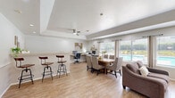 Located at 7401 Phoenix Ave in El Paso, TX, this single-floor community space offers a thoughtfully designed open-plan layout that seamlessly blends multiple functional areas into one cohesive environment. The interior is finished with warm wood-look flooring throughout, crisp white walls accented by a chair-rail detail, and a tray ceiling overhead — all contributing to a bright, clean aesthetic.

Upon entry, a welcoming foyer greets visitors with recessed lighting, a decorative console, and a plush area rug. A glazed French door connects this entry to the main living area. The open living room is the heart of the space, anchored by a classic white fireplace with a wall-mounted television above, flanked by large windows that frame views of an outdoor pool, allowing abundant natural light to fill the room. Comfortable seating arrangements invite relaxation.

Adjacent to the lounge, a dining area accommodates a full-sized wooden table with upholstered chairs, ideal for group gatherings. A built-in bar counter with industrial-style bar stools provides an additional casual seating option. A dedicated workstation area along the wall is equipped with desks and computers, adding a practical, functional dimension to the space. A ceiling fan with a modern profile serves the main area efficiently. The layout is open and versatile, making it well-suited for community use.