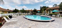 This outdoor pool area offers a welcoming and well-maintained recreational space designed for relaxation and leisure. At its center is a generously sized oval swimming pool with clear, blue-green water, accented by a classic dark tile border and equipped with entry steps and safety rails for easy access. A depth marker is visible, providing practical information for swimmers.

Surrounding the pool is a broad, level deck finished in interlocking stone pavers in a varied neutral palette, providing ample room for lounging and movement. Multiple white reclining lounge chairs are positioned around the pool's perimeter, offering comfortable spots to enjoy the open sky above. A white perimeter fence encloses the area, providing both safety and a defined boundary for the space.

Toward the back of the pool area, a shaded seating zone features a teal patio umbrella over a table and additional chairs, ideal for those seeking relief from the sun. Lush mature trees and well-maintained greenery frame the background, creating a natural, serene atmosphere. The surrounding buildings are visible beyond the fence, giving the space a community-oriented feel. The overall ambiance is bright, open, and inviting, well-suited for outdoor recreation and social gatherings.