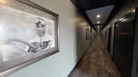 This thoughtfully designed single-floor facility offers a warm and polished environment that balances comfort with visual interest. The grand entrance foyer greets visitors with soaring ceilings, large dark-framed glass doors, and an open layout that seamlessly connects to the main lounge areas. Natural light flows generously throughout, enhancing the bright, neutral color palette of whites and soft grays.

The central lounge is an inviting gathering space furnished with cream-toned sofas, wingback chairs, and accent pillows in muted aqua and gold tones. Elegant drum chandeliers illuminate the room, while black-framed French doors add architectural definition and provide access to adjacent spaces and outdoor areas. A striking stone fireplace with a rustic wood mantel serves as a focal point in one of the seating areas, creating a cozy ambiance complemented by plush lounge chairs and tasteful décor.

A long, well-lit corridor extends through the facility, its dark trim and recessed lighting creating a refined aesthetic. The hallway is lined with framed black-and-white vintage photographs, lending the space a distinctive character and sense of history. Throughout, the interiors reflect a careful attention to detail, blending traditional warmth with contemporary elegance to create a comfortable and memorable environment.