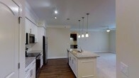 This two-story residence showcases a thoughtfully designed open-concept layout that seamlessly connects the kitchen and living areas. The kitchen is a standout feature, offering white shaker-style cabinetry, granite countertops, and a central island complete with a double basin sink. Stainless steel appliances — including a refrigerator, range, and built-in microwave — provide a sleek, modern aesthetic. A trio of pendant lights hang above the island, adding both functional illumination and a stylish focal point. Crown molding runs along the upper edges of the walls, lending an elevated, finished look to the interior. The flooring transitions from rich dark hardwood in the kitchen and dining area to plush carpet in the adjoining living space, clearly delineating each zone while maintaining a cohesive flow. Recessed and track lighting provide ample illumination throughout. A ceiling fan is visible in the living area, contributing to comfort and air circulation. The open sightlines between rooms create a sense of spaciousness and connectivity across the main floor, while the second floor offers additional living space. Overall, the interior presents a clean, contemporary aesthetic with quality finishes that balance practicality and style.