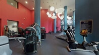 This fitness center makes a striking visual impression with its bold color palette of vivid red walls and teal cylindrical columns, creating an energetic atmosphere conducive to exercise. The space benefits from soaring double-height ceilings, which lend an open, airy quality to the interior and are enhanced by clusters of large, globe-shaped pendant lights that provide warm, even illumination throughout.

The main workout area is generously sized and houses an extensive array of strength-training machines, including Matrix and True brand equipment, free weights, and benches arranged to allow comfortable movement between stations. A large mirrored wall with a vibrant abstract mural amplifies the sense of space and adds an artistic focal point. The cardio section features a dedicated row of True brand treadmills and elliptical machines positioned alongside large windows, offering natural light and views of lush outdoor greenery. A separate cycling area accommodates several stationary bikes, also positioned near floor-to-ceiling windows that frame outdoor foliage. Durable rubber flooring throughout ensures safety and functionality. The overall ambiance is dynamic, modern, and well-appointed.