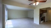 This single-floor space features an open and airy living area designed to maximize comfort and functionality. The room is defined by its vaulted ceiling, which adds a sense of height and spaciousness that extends throughout the main living zone. A ceiling fan with a light fixture is centrally mounted, providing both ventilation and ambient lighting.

The layout is notably open, with a stepped ceiling detail that visually separates distinct areas within the space while maintaining a connected, flowing feel. Neutral-toned carpeting runs throughout, offering a soft, cohesive foundation that complements the light-colored walls. The clean, minimalist wall finish creates a blank canvas adaptable to a variety of interior styles.

Natural light enters through windows, contributing to the bright and welcoming atmosphere. A floor vent indicates a forced-air climate control system integrated into the flooring. The overall design is straightforward and practical, with ample open floor space allowing for flexible furniture arrangement. The combination of vaulted ceilings, open sightlines, and neutral finishes gives this space a comfortable, livable quality suited to everyday use.