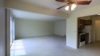 This single-floor space features an open and airy living area designed to maximize comfort and functionality. The room is defined by its vaulted ceiling, which adds a sense of height and spaciousness that extends throughout the main living zone. A ceiling fan with a light fixture is centrally mounted, providing both ventilation and ambient lighting.

The layout is notably open, with a stepped ceiling detail that visually separates distinct areas within the space while maintaining a connected, flowing feel. Neutral-toned carpeting runs throughout, offering a soft, cohesive foundation that complements the light-colored walls. The clean, minimalist wall finish creates a blank canvas adaptable to a variety of interior styles.

Natural light enters through windows, contributing to the bright and welcoming atmosphere. A floor vent indicates a forced-air climate control system integrated into the flooring. The overall design is straightforward and practical, with ample open floor space allowing for flexible furniture arrangement. The combination of vaulted ceilings, open sightlines, and neutral finishes gives this space a comfortable, livable quality suited to everyday use.