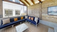 This single-level outdoor amenity space is defined by its striking architectural use of heavy timber construction, with substantial wood posts and beams forming a series of pergolas and a gabled covered pavilion. The composite decking throughout unifies the various zones while providing a low-maintenance, durable surface underfoot.

The covered pavilion serves as the centerpiece, featuring an exposed wood-paneled vaulted ceiling supported by robust timber framing. Inside, an L-shaped sectional sofa with navy cushions and accent pillows in orange and gold tones creates an inviting lounge area, complemented by a slatted wood privacy wall that adds warmth and enclosure.

Beyond the pavilion, the open deck extends into additional seating zones, including modular outdoor furniture arranged for conversation and relaxation beneath a pergola structure. Umbrella-shaded tables and chairs are visible further along, accommodating dining and casual outdoor activity.

Landscaped planters with ornamental trees are interspersed throughout, softening the hardscape and adding natural texture. The space is surrounded by a multi-story building, creating a protected courtyard atmosphere filled with natural light while maintaining a sense of community-oriented design.