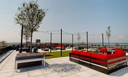 This expansive rooftop terrace offers a well-appointed outdoor amenity space with sweeping panoramic views of both a waterfront and a surrounding urban skyline. The terrace is finished with large-format light-colored pavers throughout, creating a clean, contemporary aesthetic across the entire deck.

A central artificial turf lawn serves as the activity hub, equipped with multiple cornhole boards and framed by tall poles strung with globe lights, adding ambiance for evening gatherings. Surrounding the turf area are concrete raised planters featuring trees and ornamental shrubs that introduce natural greenery to the rooftop environment.

The lounge zone features modern outdoor seating including bold red-cushioned sofas and black-and-white striped accent chairs arranged around a circular fire pit table. A separate dining and bar area includes high-top red stools and a coordinating table set. A built-in outdoor grilling station with stainless steel grills is positioned along one perimeter, ideal for entertaining.

The perimeter of the terrace is lined with sleek dark metal railings and integrated planter troughs filled with gravel and varied plantings. Unobstructed views of the waterfront extend from one side, while cityscape views with buildings and development cranes are visible from the other. A rooftop mechanical enclosure with louvered white paneling and a glass entry door provides direct access to the interior.