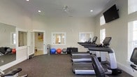 This fitness center offers a clean, functional environment designed to accommodate a variety of exercise needs. The space features high ceilings with recessed lighting and a ceiling fan, promoting air circulation and an open, airy feel. The walls are finished in a neutral tone, contributing to a bright and focused atmosphere conducive to exercise.

The flooring consists of dark rubber matting, providing durability and traction appropriate for high-traffic workout activity. A row of Life Fitness treadmills lines one side of the room, complemented by additional upright cardio machines positioned near large windows fitted with blinds. The natural light filtering through these windows enhances the overall brightness of the space.

Along the wall, several exercise balls in varying sizes—including blue, red, and grey—are neatly stored alongside workout mats, offering options for stretching and functional training. Wall-mounted mirrors reflect the equipment area, adding a sense of depth. A wall-mounted television is also present, providing entertainment during workouts. An adjacent room is visible through an open doorway, suggesting additional amenities nearby.