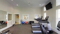 This fitness center offers a clean, functional environment designed to accommodate a variety of exercise needs. The space features high ceilings with recessed lighting and a ceiling fan, promoting air circulation and an open, airy feel. The walls are finished in a neutral tone, contributing to a bright and focused atmosphere conducive to exercise.

The flooring consists of dark rubber matting, providing durability and traction appropriate for high-traffic workout activity. A row of Life Fitness treadmills lines one side of the room, complemented by additional upright cardio machines positioned near large windows fitted with blinds. The natural light filtering through these windows enhances the overall brightness of the space.

Along the wall, several exercise balls in varying sizes—including blue, red, and grey—are neatly stored alongside workout mats, offering options for stretching and functional training. Wall-mounted mirrors reflect the equipment area, adding a sense of depth. A wall-mounted television is also present, providing entertainment during workouts. An adjacent room is visible through an open doorway, suggesting additional amenities nearby.