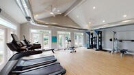 Located at 5418 Sirius Dr in Wilmington, NC, this fitness center is a thoughtfully designed single-floor facility built to support a full range of workout routines. The space is defined by soaring vaulted ceilings with exposed industrial-style HVAC ductwork, lending an open, airy atmosphere complemented by recessed lighting and dual ceiling fans for comfort during workouts. Light hardwood flooring runs throughout, adding warmth to the otherwise modern, neutral-toned interior. Natural light pours in generously through multiple sets of French doors and large windows along the perimeter, offering views of the surrounding greenery and community exterior. The main workout area is spacious and well-organized, housing a robust selection of cardio equipment including treadmills and a rowing machine, alongside a wall-mounted television for entertainment. A dedicated strength training zone features free weights, a dumbbell rack, kettlebells, TRX suspension trainers, squat racks with benches, and a cable machine system. Full-length mirrors along one wall enhance the sense of space and allow for form monitoring. A separate alcove accommodates additional strength and resistance machines. Practical amenities include a dual-level water fountain with bottle-filling station, and restroom access. The overall ambiance is bright, clean, and motivating.