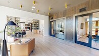 This thoughtfully designed single-floor amenity space offers a generous and open layout across a single level. The entrance welcomes visitors with light wood-toned tile flooring, gold cylindrical accent lighting, and a striking textured wood accent wall flanking glass-paneled doors that lead to private meeting rooms. The open living area is a standout, featuring floor-to-ceiling black-framed windows that flood the space with abundant natural light and frame views of the surrounding outdoor landscape and mature trees. Decorative concrete columns adorned with intricate laser-cut wood screens serve as architectural focal points, dividing the space with elegance. The living lounge is furnished with plush boucle seating, a round coffee table, and a statement brass pendant light, complemented by built-in booth-style seating with rich cognac upholstery along one wall. A wet bar with open shelving adds functional appeal. Two private dining rooms offer intimate settings, each featuring a round table, modern pendant globe lighting, and patterned area rugs. One room is equipped with a wall-mounted display screen and built-in cabinetry, ideal for meetings or private gatherings. Throughout, the cohesive palette of warm wood tones, muted neutrals, and brass accents creates a refined yet inviting contemporary ambiance.