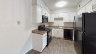 Located at 4990 Old Spartanburg Rd in Taylors, South Carolina, this single-floor unit offers a practical and comfortable layout designed for everyday living. The kitchen is a standout feature, showcasing crisp white cabinetry that extends to the ceiling for generous storage, dark granite-style countertops, and a full suite of black appliances including a refrigerator, electric range with over-the-range microwave, and dishwasher. Wood-look vinyl plank flooring runs throughout the kitchen, adding warmth and durability to the space. The open layout, visible in the dollhouse view, connects the kitchen to a central island and flows into the living area, which benefits from natural light streaming in through large windows. The living space features neutral walls and transitions to carpeted flooring, creating a cozy atmosphere. The bedroom is comfortably sized with soft carpet, neutral paint tones, and ceiling ventilation for climate control. The bathroom is clean and functional, featuring a full soaking tub with an overhead shower, curved curtain rod, wood-look vinyl flooring, and brushed nickel hardware and fixtures. Throughout the unit, a consistent neutral color palette, textured ceilings, and thoughtful finishes contribute to a cohesive and move-in-ready environment.