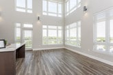 This two-story space is defined by its striking architectural design, most notably the dramatic double-height main area where towering walls of windows flood the interior with natural light and provide expansive outdoor views. The main level features warm hardwood-style flooring throughout the living and kitchen areas. The contemporary kitchen is outfitted with dark cabinetry, a white subway tile backsplash, black cooktop, stainless steel appliances including a built-in double oven and dishwasher, and light countertops that contrast cleanly against the darker finishes.

The living area benefits from oversized windows fitted with white horizontal blinds, framing views of the surrounding streetscape. A central island with a light countertop and dark base anchors the open main floor. Bathrooms are finished with large-format white tile flooring, dark vanity cabinetry, and open shelving for storage.

The upper level is accessible via a staircase and features a glass-panel railing that overlooks the main floor below. Carpeted bedroom spaces on this level are bright and well-proportioned, each with windows offering outdoor views, ample closet space, and a clean, neutral palette throughout.