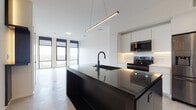 This single-floor unit at 469 Literary Rd in Cleveland's Ohio City neighborhood presents a clean, contemporary design throughout. The open-concept living and kitchen area is anchored by a striking dark-finished island with granite countertops and an integrated undermount sink with a matte black faucet. A suspended linear pendant light above the island adds a refined touch. The kitchen is equipped with stainless steel appliances, including a French door refrigerator, electric range, and built-in microwave, all complemented by white flat-front cabinetry and a subtle tile backsplash. Light wood-tone flooring runs throughout the main living areas, enhancing the bright, airy atmosphere created by large floor-to-ceiling windows that frame expansive neighborhood views. The bedroom is generously proportioned with the same light flooring and access to a walk-in closet. The bathroom features a wall-mounted floating vanity, a backlit round mirror, dark tile flooring, and a walk-in shower with white textured tile surround and black-framed glass door. Additional highlights include a stacked in-unit washer and dryer, built-in open shelving for storage, and a clean, minimalist aesthetic throughout.