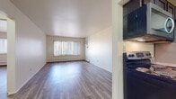 Located at 300 E Malcolm X St in Lansing, Michigan, this single-floor unit offers a clean, functional layout with consistent modern finishes throughout. The open living area is bright and welcoming, featuring durable luxury vinyl plank flooring in a warm wood tone that extends seamlessly through the space. Large windows dressed with vertical blinds allow generous natural light to fill the room while providing outdoor views of the surrounding landscape. A wall-mounted air conditioning unit and baseboard heating ensure year-round comfort.

The galley-style kitchen is efficiently designed with rich dark cabinetry, granite-style countertops, and stainless steel appliances including a refrigerator, electric range, built-in microwave, and a double sink — providing ample functionality for everyday cooking.

The entryway, accented by a solid wood door, opens into a central corridor connecting the main living areas. The layout, as seen in the dollhouse view, clearly defines distinct zones for living, cooking, and sleeping.

The bathroom is finished with tile flooring, a wood-toned vanity with a white countertop, and a tiled shower-tub combination, offering a practical and well-appointed space. Overall, the unit presents a tidy, move-in-ready environment with cohesive finishes throughout.