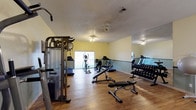 This fitness center offers a practical and well-organized workout environment designed to accommodate a range of exercise routines. The space features hardwood-style flooring throughout, providing a clean and durable surface suited for both cardio and strength training activities. Walls painted in a soft yellow hue create an energizing atmosphere, while a large mirrored wall along one side visually expands the space and allows users to monitor their form during workouts. The room is outfitted with a comprehensive selection of equipment, including a multi-station cable machine, a adjustable weight bench, a full dumbbell rack, and multiple cardio machines such as treadmills and an elliptical trainer. A sliding glass door allows natural light to filter into the space, adding to the overall brightness and openness of the room. The textured ceiling features a ceiling fan, contributing to air circulation during intense workout sessions. Smoke and ventilation systems are also visible, reflecting attention to safety and comfort. This fitness center provides a complete and functional environment for residents or members looking to maintain an active lifestyle without leaving the premises.
