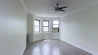 This single-floor space combines classic architectural details with functional design. The main living area is highlighted by a distinctive bay window configuration, where three double-hung windows with dark frames allow generous natural light to fill the room from multiple angles. The white-painted walls and crown molding trim contribute to a clean, polished aesthetic throughout.

Hardwood flooring in a warm gray-brown tone runs across the entire space, adding both character and durability underfoot. A five-blade ceiling fan is centrally positioned, providing efficient air circulation year-round. A traditional cast-iron radiator situated beneath the bay windows serves as both a functional heating element and a nod to the building's classic character. An additional window-unit air conditioner ensures climate control during warmer months.

The crisp white baseboards and window casings provide elegant contrast against the darker window frames and flooring, creating a cohesive visual balance. The overall ambiance is light, open, and neutral, offering a versatile backdrop well-suited to a variety of interior styles and furnishing choices.