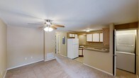 This single-floor space offers a practical and functional open-concept layout, where the living area and kitchen flow together seamlessly. The living area is finished with wall-to-wall carpeting in a warm neutral tone, creating a comfortable and understated atmosphere. A ceiling fan with a built-in light fixture is centrally mounted, providing both air circulation and ambient lighting. A floor vent indicates a forced-air climate control system throughout the space.

The kitchen is situated adjacent to the living area and is defined by a raised counter that serves as a natural dividing bar between the two spaces. Light wood cabinetry lines the upper and lower sections of the kitchen, offering ample storage. White appliances, including a full-size refrigerator and a cooktop, complement the cabinetry, while a sink with a window nearby allows for natural light. The kitchen floor transitions to tile, distinguishing it from the carpeted living area.

A hallway visible from the living area leads to additional rooms, and an electrical panel is accessible along the wall. The overall palette of soft beige and warm neutrals gives the space a cohesive, clean, and move-in-ready appearance.