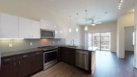 Located at 8255 Walnut Hill Lane in Dallas, Texas, this single-floor residence showcases a contemporary design with dark hardwood-style flooring flowing consistently throughout the space. The open-concept layout connects the kitchen and living area seamlessly, creating a spacious, functional environment.

The kitchen is well-appointed with dark espresso cabinetry contrasted by crisp white upper cabinets and a mosaic tile backsplash. Dark granite countertops extend across the workspace, complemented by stainless steel appliances including a range, microwave, and dishwasher. Pendant lighting hangs above the kitchen island, adding both task illumination and visual interest. A ceiling fan serves the adjacent living area, which benefits from natural light and direct access to a private balcony through sliding glass doors.

An alternate kitchen configuration, visible in additional images, features a peninsula layout with white cabinetry, a French door refrigerator, and track lighting, suggesting multiple available floor plan options within the property.

The bathroom presents a double-sink vanity with dark cabinetry, granite countertops, framed mirrors, and a full soaking tub with tile surround. Bedroom spaces are bright and neutral, with large windows allowing ample natural light. Generous closet storage is evident throughout the unit.