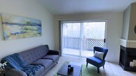 This single-floor space centers around a welcoming living area designed to maximize natural light and outdoor connectivity. The room features large sliding glass doors that open onto a private balcony enclosed with a lattice railing, framing a lush, tree-lined backdrop that brings a sense of calm and nature indoors. Vertical blinds along the sliding doors offer adjustable light control while maintaining the airy feel of the space.

The living area is finished with light-toned carpet and neutral walls, creating a versatile backdrop for furnishings. A tufted mid-century style sofa and a coordinating accent chair in navy blue anchor the seating arrangement, complemented by a low-profile wooden coffee table and a green area rug. A landscape-inspired artwork on the wall adds a touch of color and reinforces the nature-focused aesthetic of the room.

The overall ambiance is relaxed and functional, with the interplay of natural light from the balcony doors lending the interior a bright and open character throughout the day. The layout feels cohesive and thoughtfully arranged, offering a comfortable living environment with pleasant outdoor views.