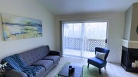 This single-floor space centers around a welcoming living area designed to maximize natural light and outdoor connectivity. The room features large sliding glass doors that open onto a private balcony enclosed with a lattice railing, framing a lush, tree-lined backdrop that brings a sense of calm and nature indoors. Vertical blinds along the sliding doors offer adjustable light control while maintaining the airy feel of the space.

The living area is finished with light-toned carpet and neutral walls, creating a versatile backdrop for furnishings. A tufted mid-century style sofa and a coordinating accent chair in navy blue anchor the seating arrangement, complemented by a low-profile wooden coffee table and a green area rug. A landscape-inspired artwork on the wall adds a touch of color and reinforces the nature-focused aesthetic of the room.

The overall ambiance is relaxed and functional, with the interplay of natural light from the balcony doors lending the interior a bright and open character throughout the day. The layout feels cohesive and thoughtfully arranged, offering a comfortable living environment with pleasant outdoor views.