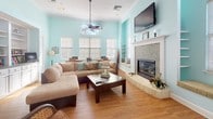 This single-floor community space exudes a relaxed, coastal charm through its consistent palette of soft aqua walls, warm hardwood-style flooring, and crisp white trim. The open layout connects several distinct functional zones that flow naturally into one another.

The living area serves as the heart of the space, anchored by a tiled fireplace with a white mantle and a wall-mounted flat-screen television. A large sectional sofa and a ceiling fan with pendant light create a comfortable, inviting atmosphere. Tall windows with horizontal blinds allow generous natural light to fill the room, while a built-in window seat offers additional seating with views of the outdoors.

Adjacent to the living area, a designated workspace features a wooden desk with a computer workstation, ideal for productivity. Open wall shelving provides practical storage throughout this zone.

The dining area accommodates a round wooden table with white-framed chairs, complemented by a built-in nook with open shelves and a cushioned bench seat. Tall windows and transom-style upper windows draw in abundant daylight.

A kitchenette rounds out the space, equipped with white cabinetry, a sink, a water dispenser, and counter space for convenient food and beverage service. French doors visible throughout the space enhance the airy, open feel and connect areas seamlessly.