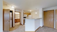 This single-floor residential unit offers a thoughtfully designed open-concept layout that maximizes functionality and flow. The kitchen serves as the heart of the space, featuring warm honey-toned wood cabinetry, a full suite of white appliances including a refrigerator, range, and built-in microwave, and a peninsula breakfast bar that provides additional counter space and separates the kitchen from the main living area. A pendant light fixture hangs above the peninsula, adding a subtle decorative touch. The living area is finished with plush carpeting in a neutral tone, creating a comfortable and cohesive atmosphere throughout. Just off the main living space, a bathroom is visible, outfitted with a well-lit vanity featuring matching wood cabinetry and a sink, offering a consistent design language that ties the unit together. The neutral white and beige color palette throughout the space lends a clean, bright, and welcoming ambiance. Flush-mount ceiling lighting ensures even illumination throughout the unit. The overall layout is efficient and practical, making excellent use of the available footprint while maintaining an open and airy feel.