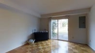 This two-story home offers a straightforward and functional layout defined by classic architectural details and practical design. The main level features distinctive parquet hardwood flooring in a warm checkerboard pattern, adding a timeless character to the space. A central hallway connects the entry area to the main living room, where natural light pours in abundantly through a large sliding glass door. This door serves as both a visual focal point and a direct access point to the outdoor greenery beyond, offering pleasant views of a landscaped yard with mature trees and shrubs.

The living area is generously proportioned, with neutral white walls and standard ceiling heights that create a clean, open atmosphere. The sliding door is framed by floor-length curtains, allowing for flexible light control throughout the day.

A staircase with dark-stained wooden treads and a light oak handrail connects the main floor to the upper level. The stairwell is well-lit, with white walls lending a bright transition between floors. The overall palette of the home is neutral and understated, providing a versatile backdrop suitable for a variety of interior styles.