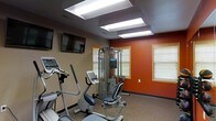 This fitness room is designed to support a well-rounded exercise routine in a compact, functional space. The room is illuminated by a series of recessed rectangular fluorescent light panels set into the ceiling, ensuring bright, even lighting throughout. Two wall-mounted flat-screen televisions are positioned for easy viewing during workouts, adding an entertainment element to exercise sessions.

The equipment selection includes multiple cardio machines — among them an elliptical trainer and a stationary bike — as well as a multi-station cable resistance machine that supports a variety of strength training exercises. The flooring features a durable, speckled rubberized surface appropriate for fitness use and easy maintenance.

The walls display a two-tone color scheme, with a warm terracotta-red accent wall contrasting against a neutral beige on the remaining walls, creating an energizing atmosphere. Wood-trimmed windows with horizontal blinds allow for natural light while maintaining privacy. The overall design is practical and straightforward, providing residents or guests with the essential tools needed for both cardiovascular and strength-based workouts in a clean, well-lit environment.