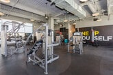 Located at 1903 E First St in Santa Ana, California, this single-floor fitness center offers a well-equipped, industrial-modern workout environment designed to accommodate a wide range of training styles. The space features exposed concrete ceilings with visible ductwork and structural elements, accented by warm pendant lighting that creates an energetic yet approachable atmosphere. Dark rubber flooring runs throughout, providing durability and traction across all training zones.

The main workout floor is generously proportioned, housing an extensive selection of SportsArt and Cybex cardio machines, including treadmills, ellipticals, stationary bikes, and recumbent cycles. A dedicated strength training area includes cable machines, multi-functional rack systems, a full dumbbell rack, and adjustable benches. Bold motivational graphics — including a prominent "ONE UP YOURSELF" mural — energize the space visually.

A standout feature is the cardio alcove enclosed by a full-panel glass garage door, which opens to outdoor views of palm trees and the surrounding landscape, flooding the area with natural light. Large windows along the perimeter further enhance brightness throughout the facility. A separate strength zone with a power rack and free weight storage offers a focused environment for heavier training. The overall design is sleek, functional, and purpose-built for serious fitness use.