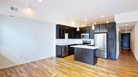 This single-floor space showcases a contemporary open-concept layout centered around a well-appointed kitchen. Dark espresso cabinetry lines the walls, providing a striking contrast against the light countertops and white walls. The kitchen is fully equipped with stainless steel appliances, including a French door refrigerator, electric range, built-in microwave, and dishwasher. A central island with a light countertop serves as both a prep area and casual gathering space, enhancing the kitchen's functionality. Overhead, a combination of track lighting and pendant lights illuminates the space with warm, focused light, adding a stylish, modern touch. The flooring throughout features warm-toned hardwood, lending a natural and inviting quality to the interior. The open layout between the kitchen and the adjacent living area creates a seamless flow, making the space feel expansive and versatile. Clean white walls and smooth ceilings contribute to the bright, airy ambiance, while the minimalist design aesthetic ensures a timeless and practical living environment.
