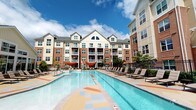 This outdoor amenity space is centered around a generously sized swimming pool with clear, blue water, set against the backdrop of an attractive multi-story residential building featuring a blend of brick and light-colored siding with gabled rooflines and private balconies. The pool deck is a notable design feature, finished in a distinctive, colorful patterned surface incorporating terracotta, blue, and sandy tones arranged in a geometric mosaic pattern, adding visual interest to the outdoor environment. Rows of lounge chairs line the poolside, offering ample space for relaxation under open skies. Shade structures are positioned near the pool's edge, providing covered retreats on sunny days. The area is enclosed by a wrought iron fence, enhancing both security and aesthetic appeal, while mature landscaping with trees and greenery softens the hardscape surroundings. A covered poolside corridor connects to additional amenities, including ADA-compliant dual drinking fountains mounted on a brick base and clearly marked accessible restroom facilities. Lattice privacy screens define transitional passages within the space. The overall ambiance is bright, welcoming, and community-oriented, with thoughtful attention to accessibility and comfort throughout.