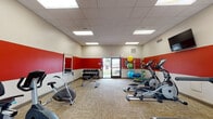 Located at 3751 Appian Way in Lexington, Kentucky, this fitness center is a well-equipped, single-floor facility designed to support a comprehensive workout routine. The space features a bold and energizing interior design, characterized by a striking red accent band running along the walls, complemented by neutral beige tones above and below. Durable, patterned carpet flooring covers the workout area, providing comfort underfoot during exercise sessions.

The facility is generously outfitted with a wide array of cardio equipment, including multiple treadmills, elliptical trainers, stationary bikes, and an air bike, allowing users to vary their cardiovascular workouts. A dedicated free weights area includes a full dumbbell rack with a mirrored wall alongside it, enabling users to monitor their form during strength training. A weight bench adds further versatility for resistance exercises.

A rack stocked with colorful exercise balls and resistance accessories provides options for flexibility and functional fitness training. A wall-mounted flat-screen television enhances the workout experience, while a water dispenser ensures convenient hydration. Natural light filters into the space through a set of large windows, offering outdoor views and creating a pleasant, well-lit environment. Recessed panel lighting in the drop ceiling ensures consistent, bright illumination throughout the entire facility.