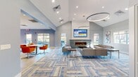 The Castlebrook Apartments clubhouse is a thoughtfully designed single-floor amenity space that combines comfort, functionality, and contemporary style. Visitors are welcomed by a textured stone accent wall bearing the Castlebrook Apartments branding, setting the tone for the modern aesthetic throughout.

The main lounge is an expansive, open area anchored by a sleek linear gas fireplace set within a concrete-finish surround, topped by a large wall-mounted flat-screen TV framed by a sculptural wave-textured panel. Soaring vaulted ceilings with recessed lighting and a striking circular pendant fixture amplify the sense of openness, while large windows and sliding glass doors invite abundant natural light and views of the outdoor patio area. Plush gray sectional seating rests on a patterned blue-and-gray area rug, creating a welcoming gathering space.

Adjacent to the lounge, a vibrant dining area features bold orange seating and white tables alongside an additional flat-screen TV set against a concrete accent wall, with colorful artwork adding visual energy to the room.

The clubhouse also includes a well-appointed kitchen equipped with stainless steel appliances, double wall ovens, light wood and white cabinetry, quartz countertops, and a stylish tile backsplash. Large windows in the kitchen offer views of the surrounding outdoor area, completing this inviting community hub.