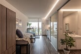 This thoughtfully designed single-floor studio offers a smart, open layout that maximizes every inch of available space. The main living and sleeping area features warm wood-tone laminate flooring that runs the full length of the unit, creating a cohesive and inviting atmosphere. Recessed LED cove lighting lines the ceiling, providing consistent, ambient illumination throughout. A floor-to-ceiling frosted glass sliding partition elegantly separates the sleeping zone from the bathroom area, adding a modern architectural touch while maintaining an open feel. The sleeping area accommodates a full-size bed with built-in storage drawers and is flanked by a tall wood-finish wardrobe, offering ample storage. Large windows at the far end of the unit flood the space with natural light and offer views to the exterior. The kitchenette is sleekly appointed with high-gloss white cabinetry, a clean white countertop, under-cabinet LED lighting, a stainless steel sink, an induction cooktop, an over-the-range microwave, and a compact refrigerator — all designed for efficient, everyday use. The bathroom is clean and minimalist, featuring wood-tone flooring consistent with the rest of the unit, a wall-mounted mirror, grab bars, and a bathtub visible through the glass partition. The overall design aesthetic is contemporary and functional.