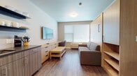 This thoughtfully designed single-floor unit offers a clean, modern aesthetic with warm wood-toned finishes and durable hardwood-style flooring throughout. The open living area is centered around a comfortable sofa and a wall-mounted flat-screen television, complemented by a streamlined storage cabinet and a low-profile coffee table. A large window with horizontal blinds fills the space with natural light while maintaining privacy. A built-in Murphy bed integrates seamlessly into the wall, maximizing the functionality of the living space without compromising its open feel.

The kitchenette is efficiently outfitted with dark laminate cabinetry, a stainless steel dishwasher, a deep sink with a modern faucet, open floating shelves for storage and display, and a smooth dark countertop with a subtle tile backsplash. The space is well-equipped for day-to-day living and meal preparation.

The bathroom features a generous vanity with wood-grain cabinetry, a wide mirror, dark countertop surface, and hardwood-style tile flooring that echoes the rest of the unit. A wall-mounted light fixture above the mirror provides bright, even illumination. The overall design of the unit balances practicality with a contemporary style, making it a highly functional and comfortable living space.