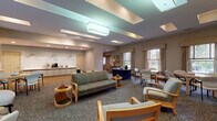 This single-floor facility is thoughtfully designed to provide both comfort and functionality across several distinct areas. The main lounge serves as the heart of the space, featuring a generous seating arrangement with upholstered sofas and armchairs positioned around small accent tables. The ceiling is a standout architectural element, with layered tray panels and large flush light fixtures that cast a soft, even glow throughout the room. Multiple windows dressed in neutral valances allow natural light to filter in, offering views of the surrounding greenery. A reading and activity corner includes built-in shelving stocked with books and decorative items, alongside a wall-mounted television and casual seating. A reception or information desk is centrally positioned within the lounge area for easy access.

The kitchen area is well-equipped, featuring an extended countertop with light gray cabinetry, pendant lighting over a beverage station, and a separate prep area with a sink, additional cabinetry, and a commercial-grade refrigerator. The coordinating blue-patterned wallpaper adds a cohesive decorative touch.

The fitness center is practical and spacious, outfitted with cardio machines, a cable weight system, and a workout bench. Exposed brick walls, a vaulted ceiling with track lighting, a ceiling fan, and large windows giving outdoor views contribute to an energizing atmosphere.