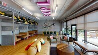 This single-floor space is a lively, travel-inspired communal lounge designed with a bold, eclectic aesthetic. The open-plan layout features warm hardwood flooring throughout, exposed ductwork along the ceiling, and abundant natural light streaming in through a full wall of floor-to-ceiling windows and glass doors that open directly onto a colorful outdoor terrace.

The interior is divided into distinct seating zones. A casual lounge area anchors one end of the space, furnished with a large tan sofa adorned with vibrant throw pillows, complemented by mismatched armchairs in earthy greens and yellows arranged around low wooden coffee tables. Nearby, a dining and collaborative work area offers long tables with green chairs, positioned beneath a striking "Field Guide to Indiana" illustrated wall mural.

Built-in open shelving lines one wall, decorated with yellow accessories, trailing plants, and small framed artworks, with booth-style seating below featuring pink hairpin legs. A games and entertainment corner houses a pool table, arcade machine, and a kitchenette marked by a "Pilot's Lounge" sign with orange cabinetry. A large pink neon sign overhead adds a dramatic focal point, reinforcing the space's playful, travel-themed character throughout.