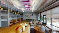 This single-floor space is a lively, travel-inspired communal lounge designed with a bold, eclectic aesthetic. The open-plan layout features warm hardwood flooring throughout, exposed ductwork along the ceiling, and abundant natural light streaming in through a full wall of floor-to-ceiling windows and glass doors that open directly onto a colorful outdoor terrace.

The interior is divided into distinct seating zones. A casual lounge area anchors one end of the space, furnished with a large tan sofa adorned with vibrant throw pillows, complemented by mismatched armchairs in earthy greens and yellows arranged around low wooden coffee tables. Nearby, a dining and collaborative work area offers long tables with green chairs, positioned beneath a striking "Field Guide to Indiana" illustrated wall mural.

Built-in open shelving lines one wall, decorated with yellow accessories, trailing plants, and small framed artworks, with booth-style seating below featuring pink hairpin legs. A games and entertainment corner houses a pool table, arcade machine, and a kitchenette marked by a "Pilot's Lounge" sign with orange cabinetry. A large pink neon sign overhead adds a dramatic focal point, reinforcing the space's playful, travel-themed character throughout.
