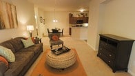 Located at 1141 Glen Iris Dr in Fayetteville, North Carolina, this single-floor residence offers a thoughtfully designed open-concept layout that seamlessly connects the living, dining, and kitchen areas into one cohesive and functional space. The living area is anchored by plush wall-to-wall carpeting in a warm neutral tone, providing a comfortable and inviting atmosphere. A decorative chandelier serves as a focal point above the dining area, adding a refined touch to the space. The kitchen features rich, dark wood cabinetry, a black appliance suite including a built-in microwave, and a peninsula counter that defines the boundary between the kitchen and living areas while offering additional prep and seating space. Recessed and flush-mount ceiling lighting throughout ensures the space is well-lit and welcoming. The open floor plan promotes a natural flow between spaces, making the layout ideal for both everyday living and entertaining. Warm beige walls complement the earthy tones found in the furnishings and accents, contributing to a cozy and harmonious interior aesthetic. A hallway visible from the main living area suggests additional private rooms beyond, completing this well-appointed single-level residence.