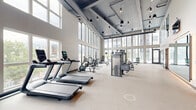 Located at The Residences at Sugar Alley in Salt Lake City, Utah, this impressive fitness and wellness center spans a generous single-floor layout designed to accommodate a full range of health and relaxation needs. The main gym area is a standout architectural achievement, featuring soaring double-height ceilings, exposed industrial ductwork, and floor-to-ceiling windows that flood the space with abundant natural light and offer sweeping views of the surrounding cityscape and mountains. The fitness floor is comprehensively outfitted with strength training equipment including free weights, cable machines, and a full rack of dumbbells, alongside dedicated cardio zones featuring treadmills, stationary bikes, and elliptical trainers. Large wall mirrors enhance the sense of openness throughout the workout area. Transitioning from the high-energy gym, the wellness side of the facility offers a serene contrast. A dedicated massage treatment room features warm, floor-to-ceiling wood slat accent walls, creating a calm and intimate atmosphere. A relaxation lounge provides cushioned chaise loungers framed by sheer white drapery panels, with large glass doors opening to outdoor views of the mountains. A utility room and support spaces round out the facility's functional offerings.
