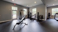 Located at 3590 Mary Adler Ave in Charleston, SC, this single-story fitness center is designed to offer a comprehensive workout experience in a clean, well-organized environment. The facility is finished throughout in neutral gray tones with crisp white trim, creating a modern and functional aesthetic.

The space is divided into distinct workout zones. The strength training area houses multiple cable machines, a weight bench, and a rack of dumbbells, all set on durable rubber flooring that extends throughout the workout areas. A dedicated cardio zone accommodates treadmills and elliptical trainers, each positioned near large windows that frame views of the surrounding greenery, providing ample natural light and an open feel during workouts. Wall-mounted mirrors in the cardio area further enhance the sense of space.

An arched entryway connects the workout zones to a transitional lounge area, where a wall-mounted television, shelving stocked with towels, and a service window create a welcoming reception point. Adjacent to this is a small carpeted lounge nook with two accent chairs and a side table, ideal for resting or waiting. Restroom facilities, including an ADA-accessible restroom, are accessible from the central corridor. Recessed lighting and multiple windows ensure the entire facility remains bright and comfortable.