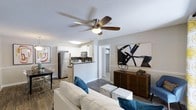 This single-floor residence showcases a thoughtfully designed open-concept layout that seamlessly connects the living area, dining space, and kitchen. The living room is a welcoming focal point, featuring wood-style flooring, neutral-toned walls, and a ceiling fan with an integrated light fixture that enhances air circulation and illumination. A comfortable seating arrangement anchors the space, complemented by contemporary artwork and stylish décor accents.

The kitchen is efficiently designed with white cabinetry, dark countertops, and a full suite of stainless steel appliances, including a refrigerator and microwave. A breakfast bar provides a natural transition between the kitchen and the living area, making it ideal for casual dining or entertaining. A dedicated dining area with seating is situated nearby, offering a distinct space for meals.

A hallway visible from the main living area leads to additional rooms, including what appears to be a bedroom with a walk-in closet, adding to the unit's storage and privacy. The textured ceilings and consistent flooring throughout contribute to a cohesive aesthetic. The overall ambiance is bright, modern, and functional, designed to maximize comfort and usability within a compact, efficient footprint.