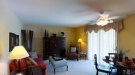This single-floor space offers a comfortable and well-appointed interior designed with a classic, homey aesthetic. The living area serves as the heart of the space, featuring warm golden-yellow walls that create an inviting atmosphere. Soft, plush carpeting runs throughout, adding to the overall sense of comfort and warmth. A ceiling fan with a light fixture is centrally positioned overhead, providing both air circulation and ambient lighting. Large windows dressed with elegant valances and sheer vertical blinds allow generous natural light to flow into the room while offering privacy. The living area is thoughtfully arranged with seating that includes a red upholstered sofa and a brown leather accent chair paired with a side table and lamp, creating a functional and cozy sitting arrangement. A substantial dark wood armoire with multiple drawers and cabinet doors serves as a prominent storage and display piece along one wall. A tufted upholstered ottoman with nail-head trim doubles as both a coffee table and additional seating. Adjacent to the living area, a glimpse of a dining space is visible, hinting at an open, connected layout ideal for everyday living and entertaining. Overall, the space presents a classic, well-maintained interior with a warm and welcoming character.