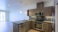 This single-floor space showcases a thoughtfully designed modern kitchen that serves as its centerpiece. The cabinetry is finished in a warm dark wood tone, offering ample storage through both upper and lower units, including an open wine rack section that adds a functional decorative touch. A dark stone countertop extends across the L-shaped layout and wraps around a peninsula, providing generous workspace and additional seating potential.

The kitchen is equipped with a full suite of stainless steel appliances, including a built-in oven, ceramic cooktop, over-the-range microwave, and a dishwasher integrated seamlessly into the cabinetry. A sleek, arched faucet sits above a stainless steel undermount sink within the peninsula. The backsplash features vertically laid dark tile in a subtle grid pattern, adding texture and visual depth to the cooking area.

Two teardrop-shaped pendant lights hang above the peninsula, providing focused task lighting while contributing to the contemporary aesthetic. Light hardwood flooring contrasts warmly against the darker cabinetry, and the bright white walls enhance the overall sense of openness and cleanliness throughout the space.