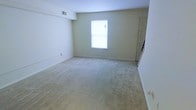 This two-floor space presents a straightforward, functional layout with a clean and neutral aesthetic throughout. The pictured room features wall-to-wall beige carpeting that extends across a generously sized open area, providing a soft and comfortable underfoot experience. The walls are finished in a light, off-white tone that contributes to a bright and airy atmosphere. A single window positioned on the far wall allows natural light to filter into the room, enhancing the overall sense of openness. A ceiling-mounted HVAC vent ensures climate control, while white baseboards frame the perimeter of the room neatly. A doorway visible to the right side of the room suggests access to additional spaces, hinting at a connected layout across the floor. The room's simplicity and uncluttered state make it highly adaptable for a variety of uses. The two-floor configuration of the overall space provides separation between living areas, offering both functionality and versatility for occupants.