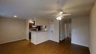 This single-floor residence offers a practical and well-appointed open-concept layout that integrates the living and kitchen areas into one cohesive space. Rich, dark hardwood-style flooring runs throughout, providing a warm and grounded aesthetic that complements the neutral wall tones.

The kitchen is positioned at the heart of the space, anchored by a peninsula with granite countertops that doubles as a breakfast bar. Dark cabinetry lines the kitchen walls, offering ample storage, while stainless steel appliances — including a microwave and range — add a modern touch. A range hood overhead ensures proper ventilation.

The living area is generously sized, illuminated by a ceiling fan with integrated lighting that enhances both airflow and ambiance. Built-in shelving is conveniently recessed into the wall, providing additional display and storage options. Visible HVAC vents are strategically placed throughout the ceiling, ensuring consistent climate control.

A hallway extends from the main living space, leading to additional rooms, suggesting a thoughtful floor plan with clearly defined private and communal zones. Overall, the space presents a clean, functional design with durable finishes suited for everyday living.