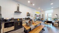Located at 144 Addison St in Boston's East Boston neighborhood, this single-floor residence features a thoughtfully designed open-concept layout that connects the living, dining, and kitchen areas into one cohesive, light-filled space. Large floor-to-ceiling doors in the living area open to a private balcony, drawing in natural light and offering views of the surrounding neighborhood. The living space is anchored by wood-tone flooring throughout and accented by a bold yellow neon sign, wire-frame shelving, and eclectic wall art that give the unit a distinctly creative character.

The kitchen is well-equipped with two-tone cabinetry—pairing light wood lower cabinets with darker upper units—dark countertops, stainless steel appliances including a full-size refrigerator, a sleek range with an industrial-style hood, and a dishwasher. Open wall-mounted shelving provides additional storage and display space. A long natural wood communal-style dining table with matching benches anchors the dining area adjacent to the kitchen.

The bedroom features a full-height pegboard accent wall that doubles as functional storage and display, a large window with roller shade offering outdoor views and ample natural light, and carpeted flooring for comfort.
