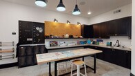 This single-floor art studio is thoughtfully designed to support a wide range of creative pursuits in a well-organized, functional environment. The space centers around a generously sized butcher-block worktable mounted on a sturdy black steel frame, its surface richly marked with layers of paint from past projects — a testament to its active use. Bar-height stools provide comfortable seating at the table, which sits atop polished concrete floors that extend throughout the room.

Along the perimeter, dark-finished cabinetry with brass hardware offers extensive storage, complemented by open shelving that displays art supplies, canvases, and finished works. A large cork board mounted above the shelving provides a practical surface for pinning references and inspiration. A black pegboard wall organizes tools and materials, while rolling metal lockers and wall-mounted paper roll holders add further utility.

Overhead, a combination of bold black industrial pendant lights and recessed ceiling fixtures ensures strong, even illumination ideal for detailed creative work. A wall-mounted television and a wooden easel round out the studio's amenities, supporting both instruction and display. A doorway connects the studio to an adjacent gathering area, enhancing the space's versatility.
