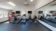 Located at 100 Bermuda Run in Statesboro, Georgia, this single-story community amenity center offers a well-rounded selection of facilities designed to support both active and social lifestyles. The fitness center is a standout feature, thoughtfully divided into distinct workout zones. One area houses a row of cardio equipment — including treadmills, ellipticals, and a stationary bike — positioned in front of floor-to-ceiling mirrors and wall-mounted televisions, with natural light streaming in through exterior glass doors. A separate strength training zone features a comprehensive dumbbell rack, multi-use cable machine, and additional resistance equipment, all set on durable rubber flooring beneath vaulted ceilings with ceiling fans. The business center provides a quiet, functional workspace with a long desk running along a deep teal-painted wall, three computer workstations, brass wall sconces, and framed abstract artwork overhead. The social lounge is an inviting open-plan space finished with wood-look flooring, bold dark trim, and a distinctive tiled bar-height island at its center. Hanging bubble chairs and colorful drapery add a playful, modern character to the room. A pool table and open seating areas complete this communal gathering space, making it ideal for relaxation and recreation.